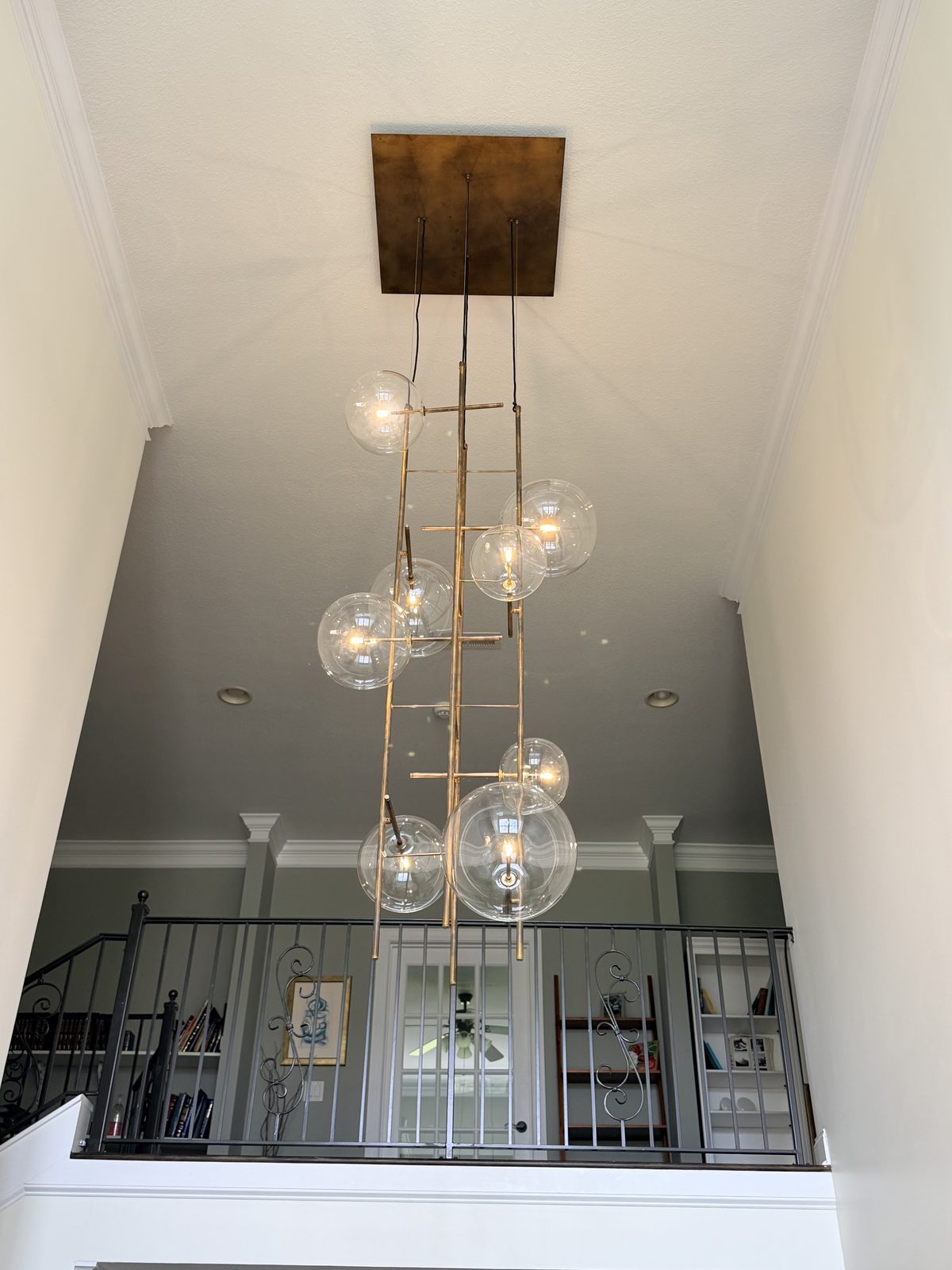 Tall brass ladder-style chandelier with clear glass globe lights hanging in a two-story foyer with crown molding and an upstairs railing