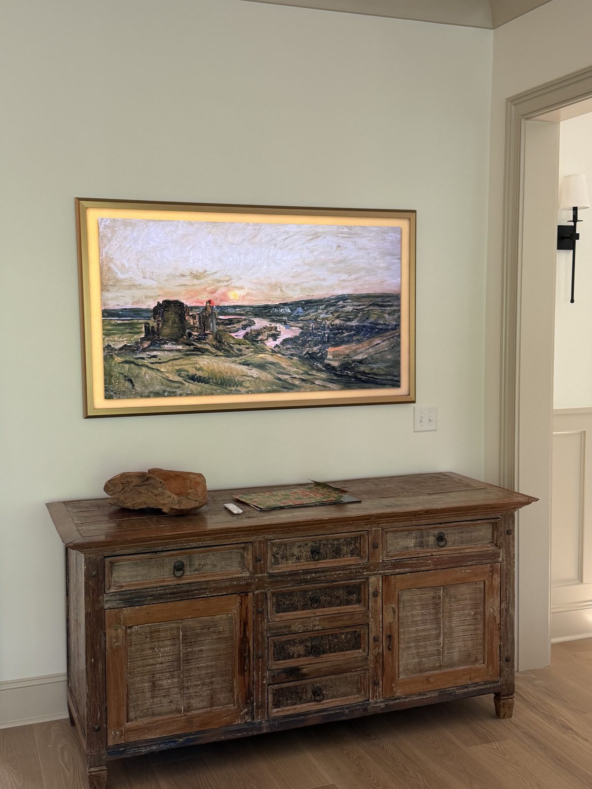 Art-style framed TV mounted flush on a soft green wall above a rustic wood sideboard, with a black wall sconce visible through a doorway