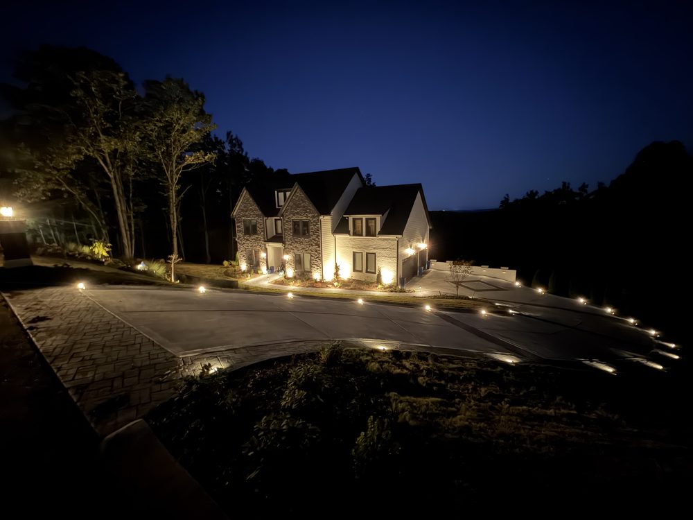 Two-story stone and siding home at dusk with an uplit facade and low path lights lining a long curved driveway