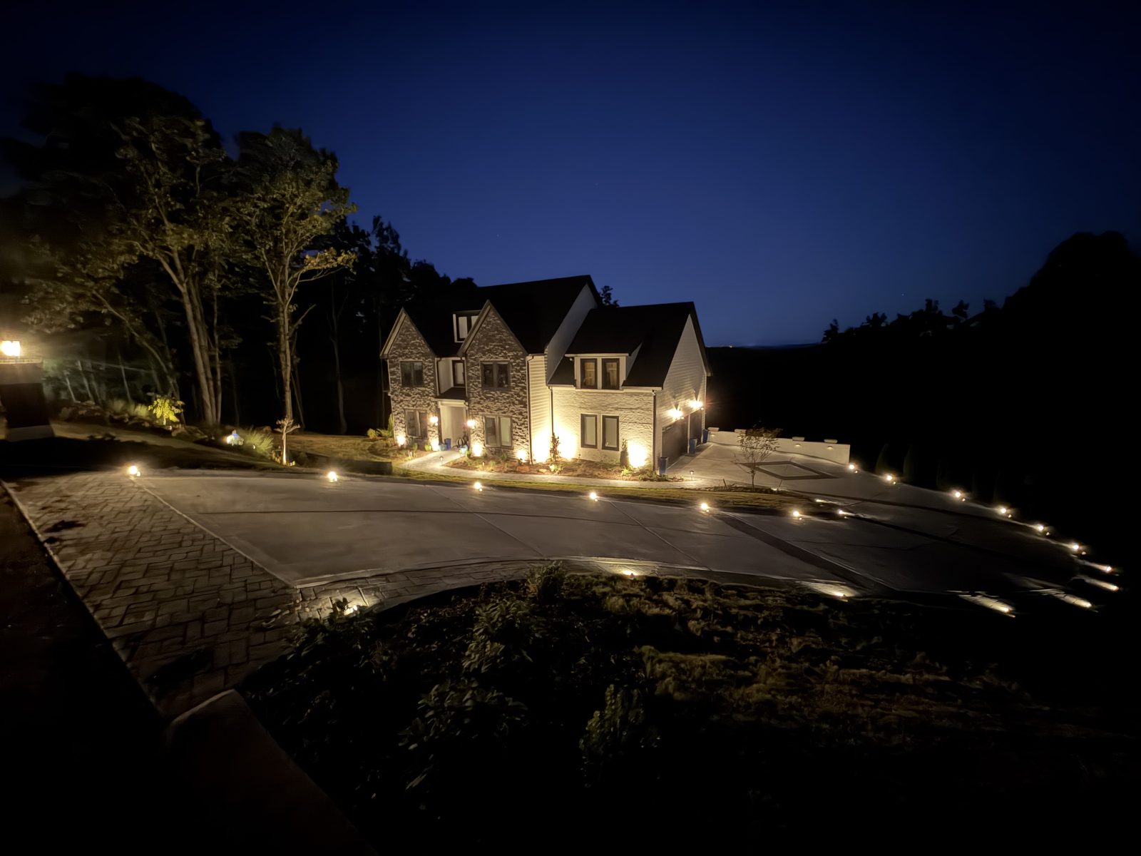Two-story stone and siding home at dusk with a softly uplit facade and a long curved driveway lined with low path lights
