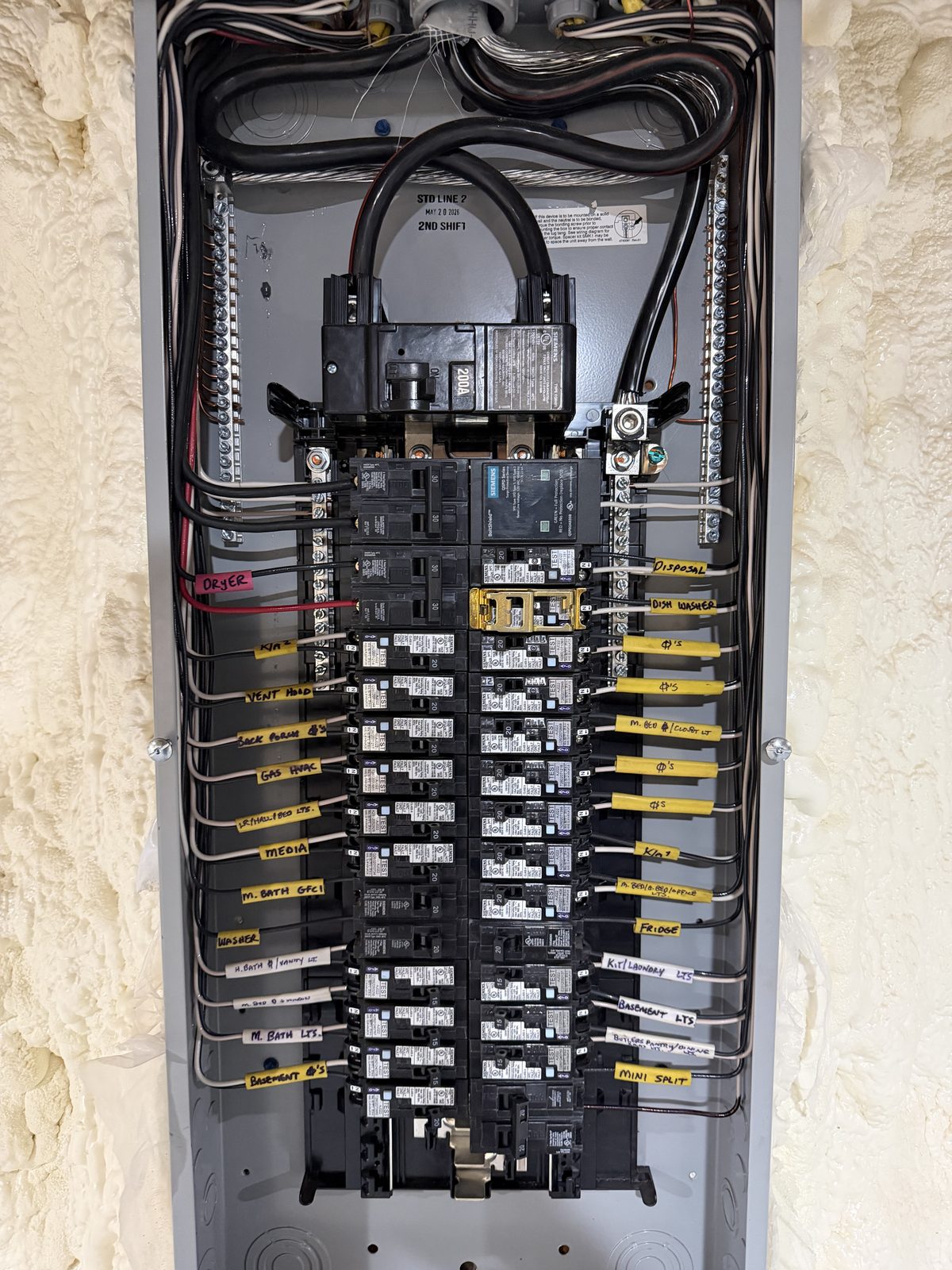 Neatly wired 200-amp residential panel with every circuit hand-labeled on yellow and white tags, set in a spray-foamed wall